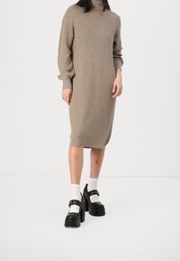 Abito in maglia a coste taupe con scollatura alta, maniche lunghe e lunghezza fino al polpaccio. Abbinato a scarpe con plateau nere e calzini bianchi.