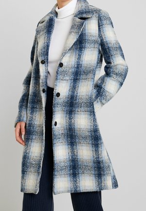 Femme portant un long manteau à carreaux bleu et blanc avec des boutons noirs sur un col roulé blanc et un pantalon sombre, main dans la poche.