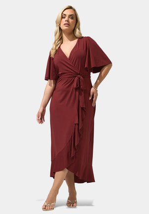 Frau in einem burgunderroten Wickelkleid mit flatternden Ärmeln, V-Ausschnitt und Taillenband, stehend mit überkreuzten Beinen und goldenen Riemchen-High Heels.
