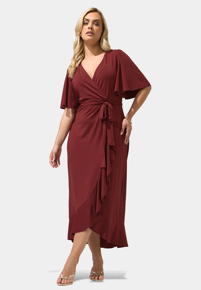 Woman wearing a burgundy wrap dress with flutter sleeves, a V-neck, and a tie waist, standing with legs crossed and gold strappy heels.