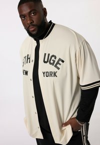 Maglietta da baseball in stile beanie color crema con scritte nere, maniche corte e dettagli a righe. Presenta chiusura con bottoni a pressione e una vestibilità rilassata.