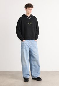 Schwarzer Kapuzenpullover mit grünem Grafikdesign auf der Brust, kombiniert mit hellblauen, lässigen Jeans und schwarzem klobigen Schuhen. Einheitlicher Hintergrund.