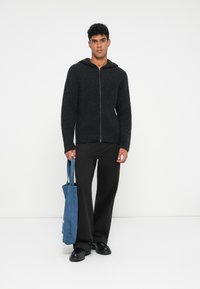 Mørk grå hettegenser med glidelås og strukturert stoff, kombinert med løse svarte bukser og en blå denim totebag, båret av en stående mannlig modell.