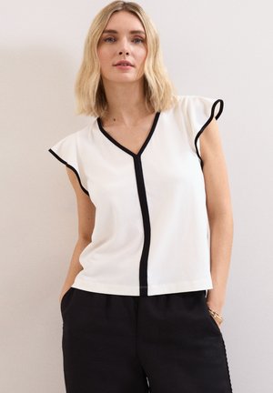 Woman with shoulder-length blonde hair wearing a white blouse with black trim and black pants, standing against a plain light background.