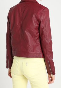 Giacca di pelle bordeaux con un design aderente, cuciture visibili e polsini con zip, abbinata a pantaloni giallo chiaro. Texture liscia.