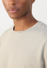Svetainės spalvos t-shirt su apvaliu kaklu ir trumpomis rankovėmis. Lygus audinio paviršius. Artimas fokusas ant apykaklės ir peties srities.