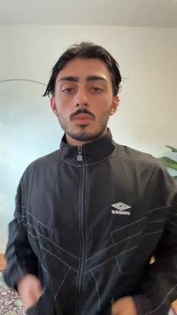 Svart jacka med dragkedja och vita detaljer och logotyp, med hög krage och kontrasterande sömnadsdetaljer. Enfärgad bakgrund med grönska.