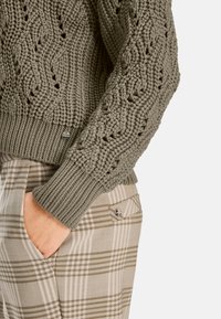 Olivgrüner Strickpullover mit strukturiertem Muster und gerippten Bündchen, kombiniert mit beigen Karohosen mit Taschen und metallischen Akzenten.