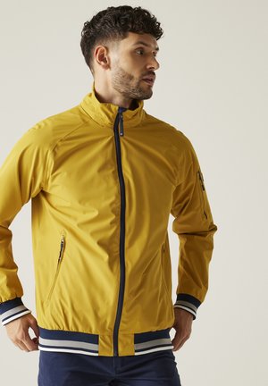 Man with curly hair wearing a yellow zip-up jacket with navy and white striped cuffs and hem, looking to the side.