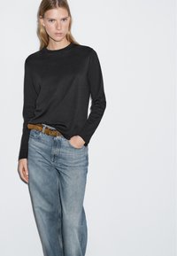 Schwarzes Langarmshirt kombiniert mit hellblauen, hochgeschnittenen Jeans und einem braunen Gürtel. Glatter Stoff mit klassischer Passform.