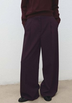 Pantalon à jambes larges d'un bordeaux profond, confectionné dans un tissu texturé. Il possède des plis à l'avant et des poches latérales, associé à des chaussures noires.