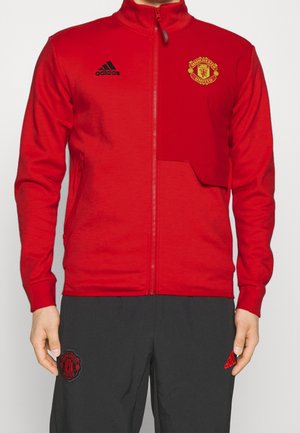 Veste rouge zippée avec écusson de Manchester United et logo Adidas, portée avec un pantalon noir arborant les logos assortis de Manchester United et Adidas.
