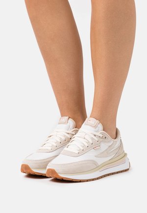 Pieds portant des baskets FILA blanches et beiges avec des lacets blancs et des semelles en caoutchouc brun, présentés sur un fond clair uni.