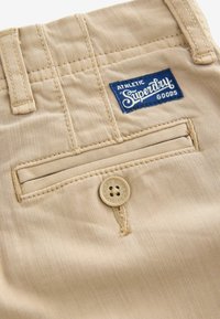 Pantaloni in cotone beige con una tasca posteriore, etichetta blu con marchio e un bottone tondo beige con logo. Tessuto liscio.