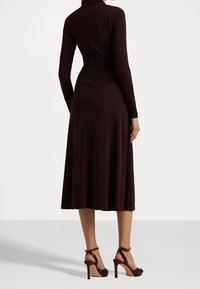 Burgundy långärmad klänning i slätt tyg, med en figursydd kropp och utsvängd kjol. Matchas med bruna högklackade sandaler.