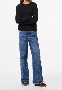 Cárdigan negro de punto con cuello redondo y cierre de botones, combinado con jeans vaqueros de pierna ancha en azul y zapatos tipo slip-on en burdeos.