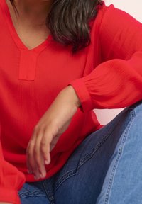 Blouse rouge à manches longues avec col en V et tissu texturé. Manches légèrement froufroutées et détail subtil à l'avant, portée avec un jean en denim.