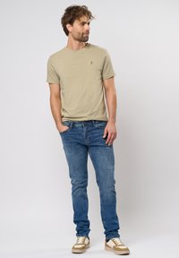 Camiseta khaki de manga corta con un pequeño logo, combinada con vaqueros azules y zapatillas marrón y blancas, de pie contra un fondo liso.