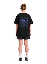 Personne avec les cheveux attachés portant un T-shirt noir oversize avec le texte "OFF DUTY GEAR" et un graphisme abstrait au dos, un short noir, des chaussettes blanches et des chaussures.