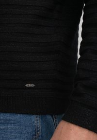 Maglione nero in tessuto strutturato con righe orizzontali, dotato di una piccola etichetta in metallo sull'orlo. Indossato con jeans in denim blu.