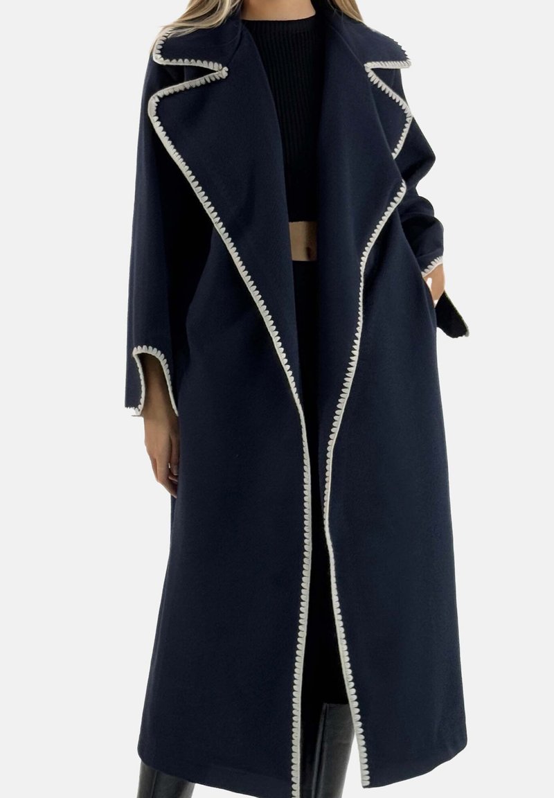 Marineblauer Mantel mit einer strukturierten Oberfläche, der weiße, gewellte Einfassungen entlang der Ränder und einen breiten Kragen aufweist. Lockerer Sitz mit langen Ärmeln.