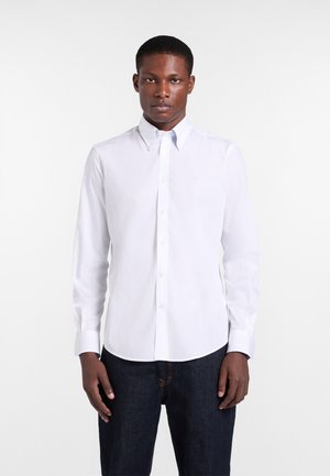 Uomo che indossa una camicia bianca aderente con bottoni e colletto, e jeans scuri, in piedi davanti a uno sfondo bianco uniforme.