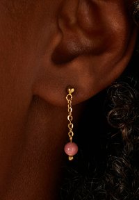 Boucles d'oreilles en chaîne dorée avec une perle rose suspendue à un petit clou. Mise au point sur l'oreille avec une texture de peau détaillée en arrière-plan.