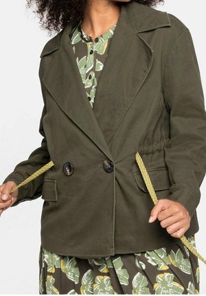 Kvinde iført olivengrøn dobbeltradet jakke over blomsterkjole, justerer gule snore ved taljen.