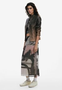 Veste bomber en bronze à motifs avec une fermeture éclair, superposée à une robe maxi fluide à imprimé abstrait ; associée à des chaussures slip-on crème.