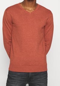 Man som bär en rostfärgad V-ringad tröja ihop med mörkgrå jeans, stående mot en enkel ljus bakgrund.