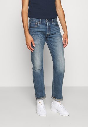 Homme portant un jean bleu délavé roulé aux chevilles, des baskets blanches et un t-shirt bleu marine foncé, debout devant un fond uni.