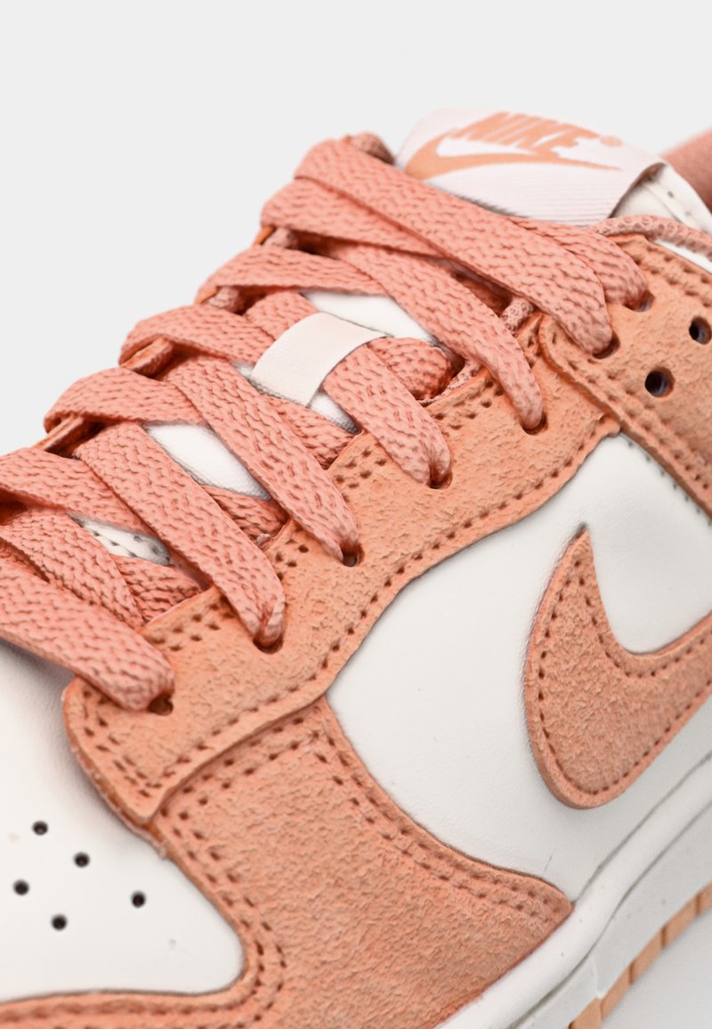 Sneaker Nike avec une base en cuir blanc, des accents en suede pêche, des lacets texturés pêche et un logo visible sur la languette.