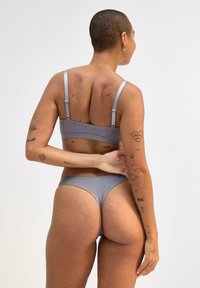 Grau Bralette- und Bikini-Set aus glattem Stoff. Das Bralette hat verstellbare Träger; der Bikini hat einen minimalen Schnitt. Dekorative Tattoos sind sichtbar.