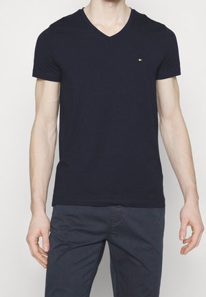 Man wearing a plain dark navy V-neck T-shirt and dark pants, standing against a plain light background.