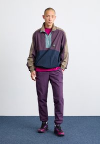 Color-blocked windbreaker in purple, navy, and green with a half-zip. Paired with matching purple pants and black sneakers with pink accents.