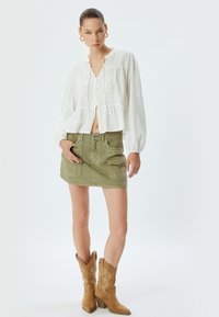 Blusa bianca cropped e texturizzata con chiusura a bottoni; mini gonna verde oliva con tasche anteriori; stivali da cowboy beige con dettagli ricamati.
