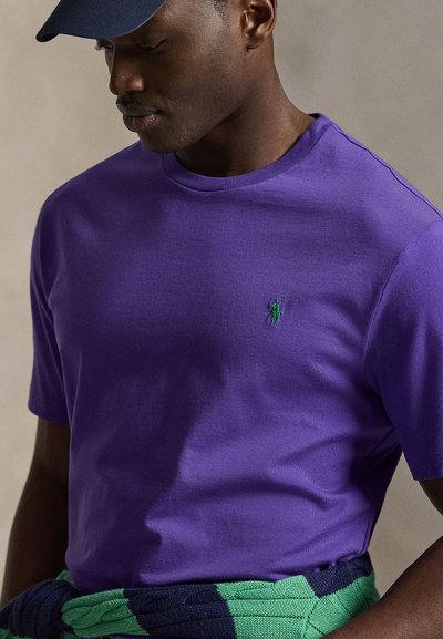 Mand iført en lilla T-shirt med grønt logo, marineblå kasket og en grøn og marineblå sweater bundet omkring hans talje.