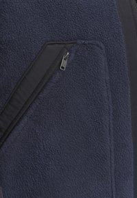 Mornarska fleece jakna s glatkim mješavinom tkanine. Ima džep na prsima sa zatvaračem i kontrastni crni panel duž linije zatvarača.