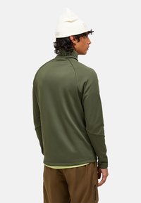 Pullover lungo a maniche lunghe verde oliva con colletto alto e texture a coste; abbinato a pantaloni marroni; berretto bianco con accentuazione del logo.