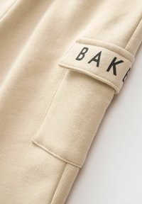 Beżowy mankiet bluzy z teksturowanej tkaniny. Przyszyta naszywka z wyraźnym napisem "BAKE" w czarnych literach. Prosty, minimalistyczny design.