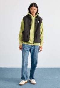 Dunkelgraue ärmellose Weste über einem grünem Tie-Dye-Hoodie, kombiniert mit hellblauen Jeans und weißen Sneakers mit sandfarbenen Akzenten.
