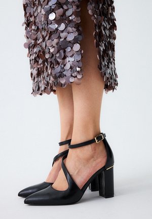 Zapatos de tacón alto de piel negra con una correa en el tobillo, que presentan puntas afiladas y un diseño recortado, sobre un vestido de lentejuelas.