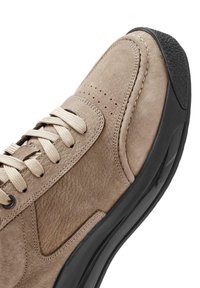 Chaussure en daim beige avec un design perforé, lacets beiges et semelle en caoutchouc noir. Elle présente une texture lisse et des détails de couture le long des bords.