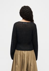 Γυναίκα με σγουρά σκούρα μαλλιά, που φοράει μαύρο αφράτο πουλόβερ με μακριά μανίκια και μπεζ πλισέ φούστα ψηλόμεση, φαίνεται από πίσω.