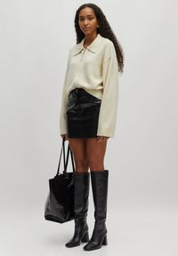 Cremfarbener Oversized-Pullover mit Kragen, Minirock aus schwarzem Krokodilmuster, kniehohe schwarze Lederstiefel und eine glänzende schwarze Tote-Tasche.
