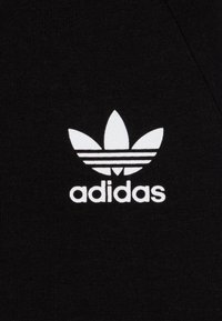 Črn bombažni material s prvim logotipom Adidas v beli barvi, s tremi horizontalnimi črtami pod logotipom in gladko teksturo.
