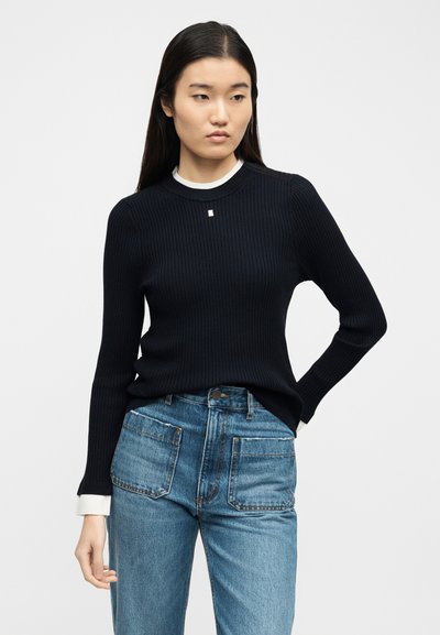 Γυναίκα με μακριά σκούρα μαλλιά που φοράει μαύρο ριγωτό πουλόβερ πάνω από άσπρο πουκάμισο και μπλε τζιν με μεγάλες μπροστινές τσέπες, στέκεται μπροστά σε απλό φόντο.