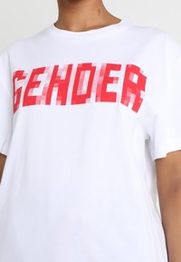 Bílý bavlněný tričko s velkým, pixelovaným červeným textem "GENDER" na přední straně. Krátké rukávy a uvolněný střih.