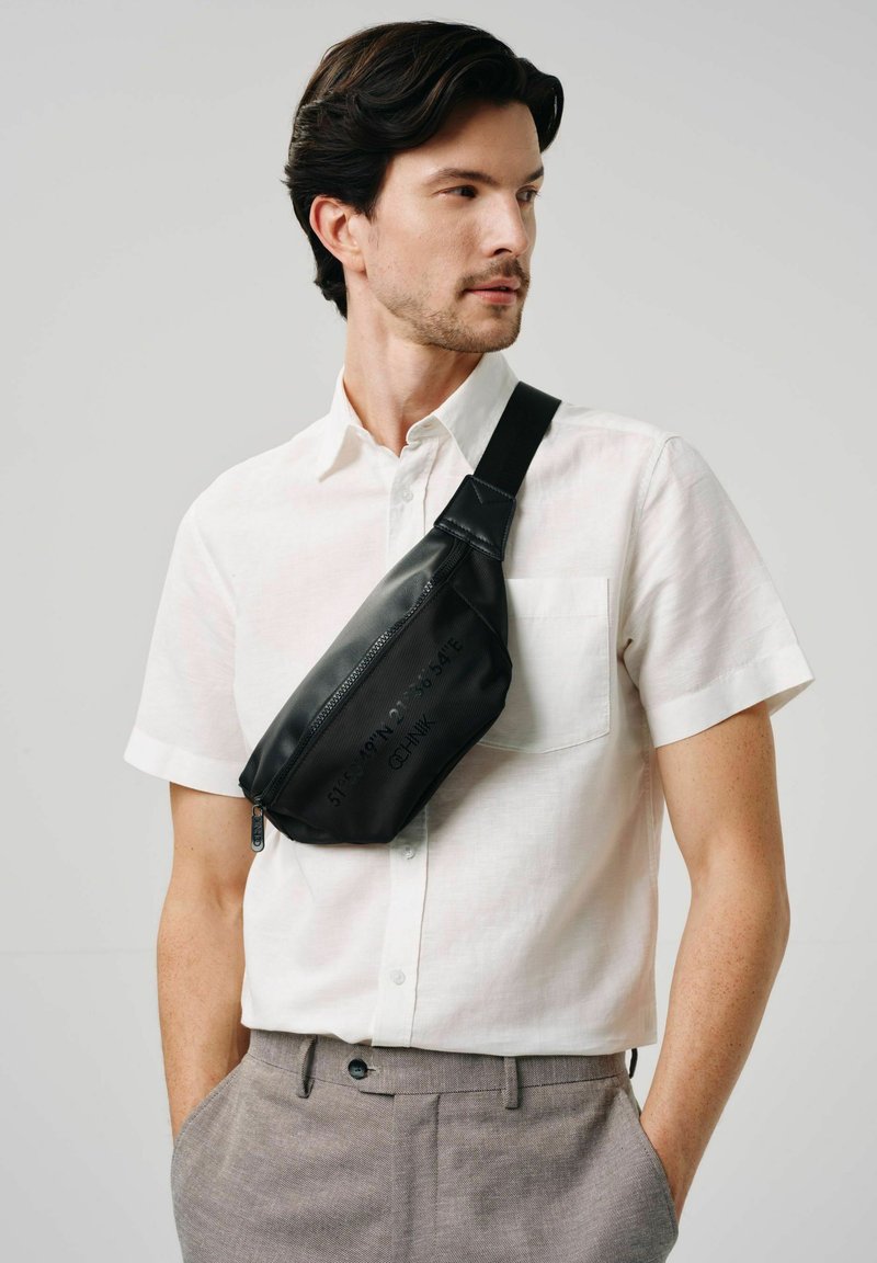 Czarna nerka o teksturowanej powierzchni i zamku błyskawicznym, noszona ukośnie na klatce piersiowej, na jasnej koszuli i szarych spodniach.