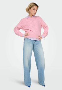 Pull rose en tricot avec un motif texturé, superposé à une chemise bleu clair à rayures, associé à un jean en denim bleu à jambe large.